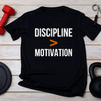 Black t-shirt with 'Discipline > Motivation' text on a wooden surface with fitness equipment.